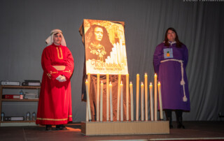 Dos jóvenes de Tobarra ataviados con túnica de tambor y de hermandad en la presentación del cartel de semana santa de Tobarra 2023, delante del cartel colocado tras una serie de velas imitando un trono.
