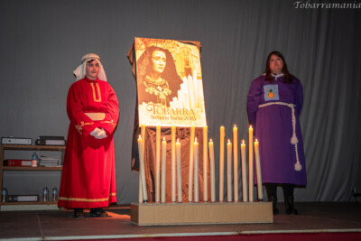 Dos jóvenes de Tobarra ataviados con túnica de tambor y de hermandad en la presentación del cartel de semana santa de Tobarra 2023, delante del cartel colocado tras una serie de velas imitando un trono.